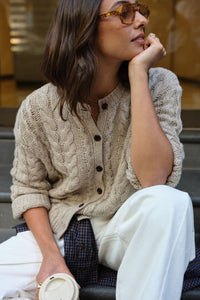 Woman wearing a beige knitted cardigan sitting on steps. CERES LIFE, ENID CABLE KNIT CARDI. 