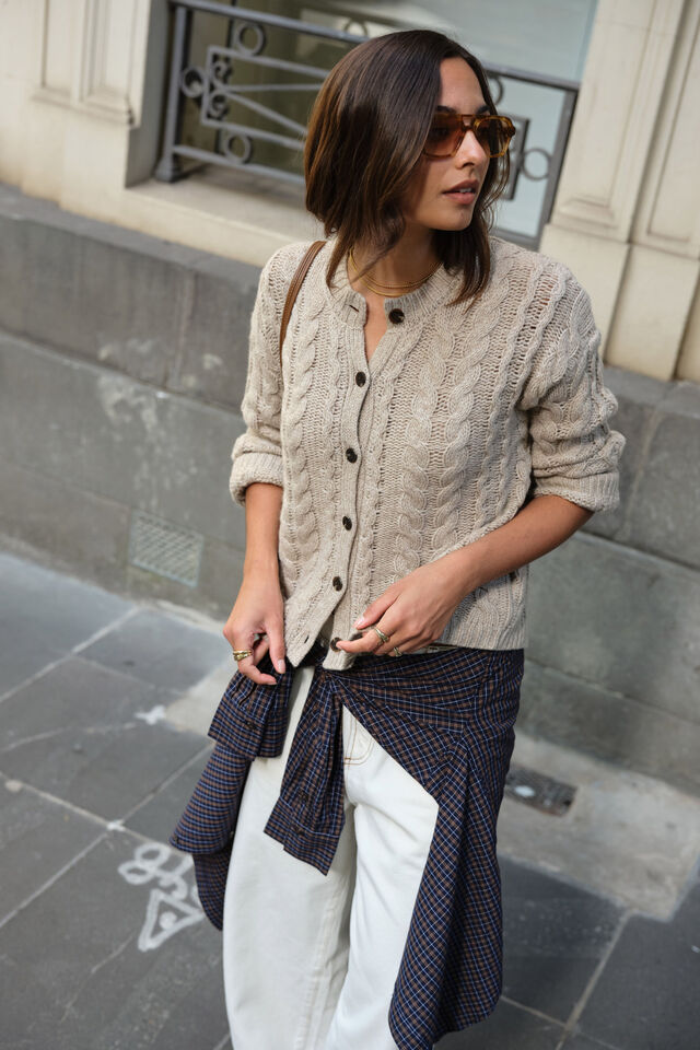 Woman wearing a beige cable knit cardigan, white pants, and a dark patterned scarf on a city street. CERES LIFE. CARDI. 