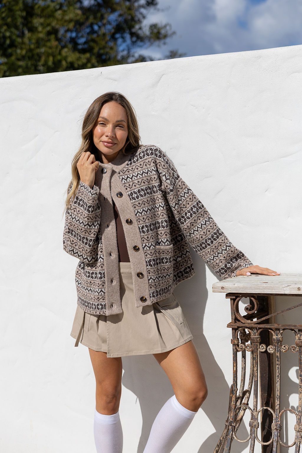 Woman wearing a patterned coat standing against a white wall with trees in the background
